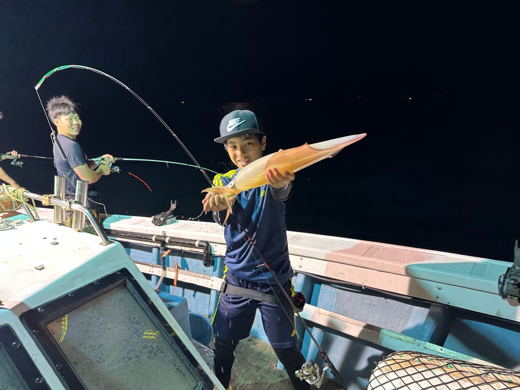 夜の船上で釣り上げた大きな白いかを持つお客さま。後ろでは竿が大きく曲がっている